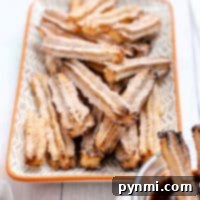 A stack of air fryer churros, filled with rich chocolate, sits on a patterned plate, with a bowl of extra chocolate sauce nearby for dipping.