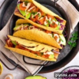 Three tacos with chicken, avocado, cabbage, and sauce sit invitingly on a plate. Beside them is a fresh avocado half and vibrant purple cabbage. These delicious tacos feature perfectly crispy taco shells made in an air fryer for the ultimate crunch experience.