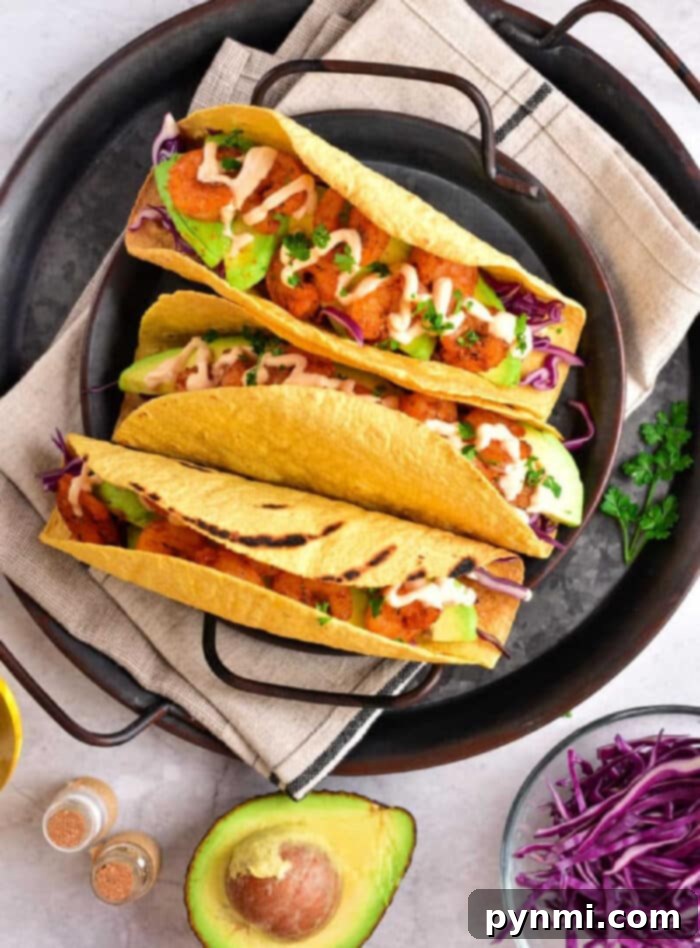 Three tacos with chicken, avocado, cabbage, and sauce sit invitingly on a plate. Beside them is a fresh avocado half and vibrant purple cabbage. These delicious tacos feature perfectly crispy taco shells made in an air fryer for the ultimate crunch experience.