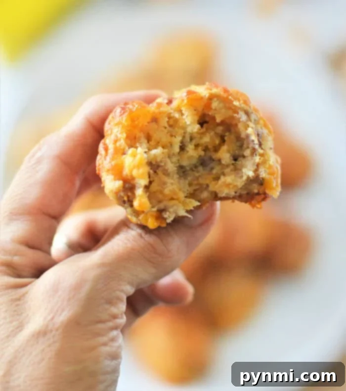 A hand holds a bitten savory Bisquick sausage balls, with a blurred background showcasing similar muffins on a plate.