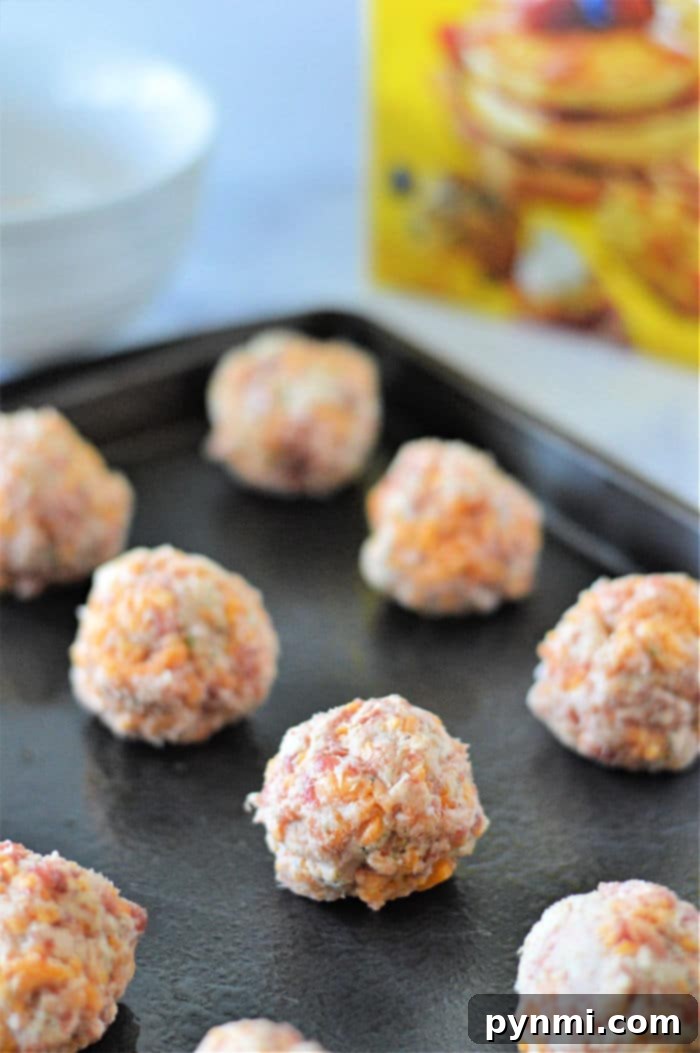 Unbaked sausage balls rest on a black baking sheet, with a cereal box subtly visible in the background, reminiscent of simplicity, much like crafting Bisquick sausage balls with just 3 ingredients.