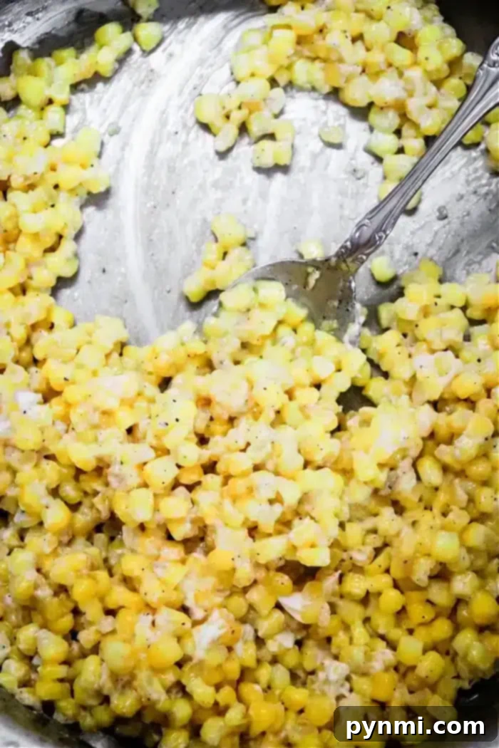 Golden Honey Butter Skillet Corn 6 A close-up of a metal dish containing honey butter skillet creamed corn. The yellow kernels are mixed with a creamy sauce, accented by black pepper, while a metal spoon rests in the dish, partially immersed in the corn mixture.
