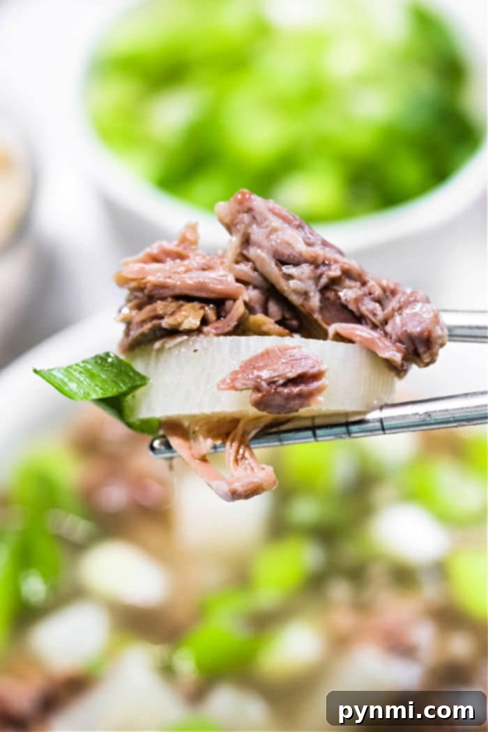 Korean Oxtail Gomtang 6 Chopsticks holding a tender piece of oxtail meat and vegetable hover above a bowl of Korean Oxtail Soup, with fresh greens adding a vibrant touch in the background.