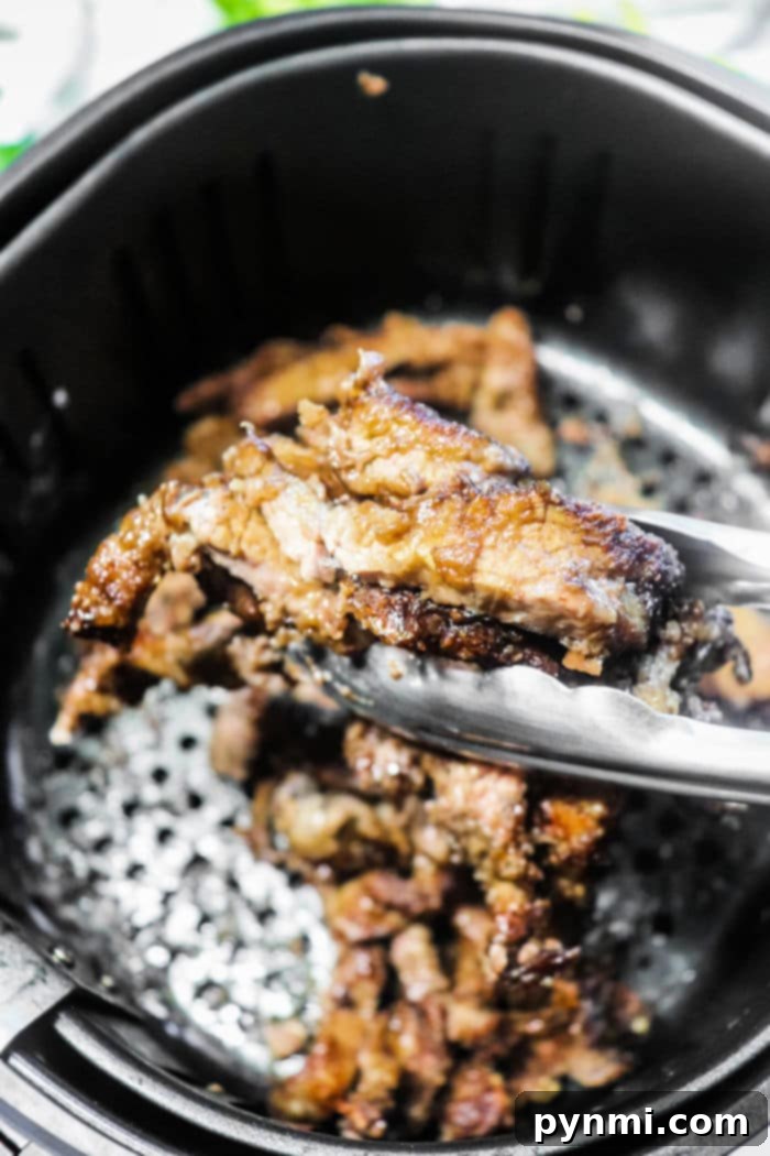 Close-up of tongs gripping perfectly cooked beef strips in an air fryer basket, showcasing the crispy delight.
