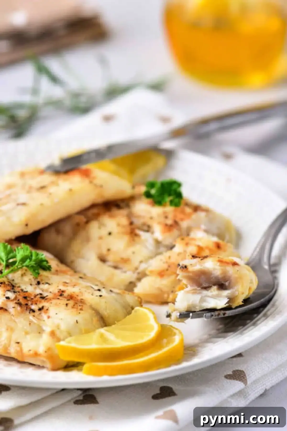 A white plate features air-fried fish fillets garnished with lemon slices and a sprig of parsley. A fork holds a piece of the flaky delight, while a blurred glass with a yellow drink sits in the background. The tablecloth is white with heart patterns, emphasizing this delicious twist on frozen cod.