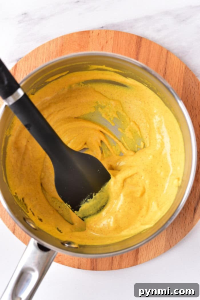 A saucepan filled with creamy hot dog mustard, being stirred by a black spatula on a wooden trivet.