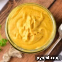 Glass jar of creamy yellow hot dog mustard on a wooden board with a spoon beside it.