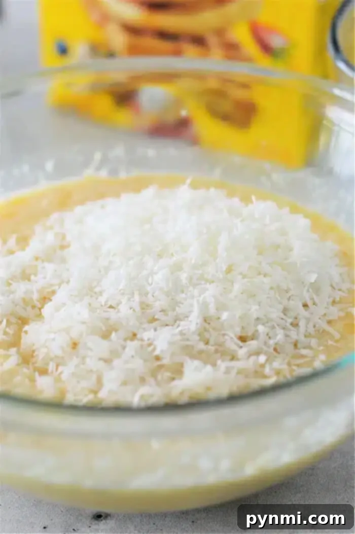 Magic Coconut Bisquick Pie 6 A glass bowl filled with the creamy Bisquick Impossible Coconut Pie batter mixture, generously topped with shredded coconut before baking. A blurry background suggests a kitchen setting with other baking ingredients.