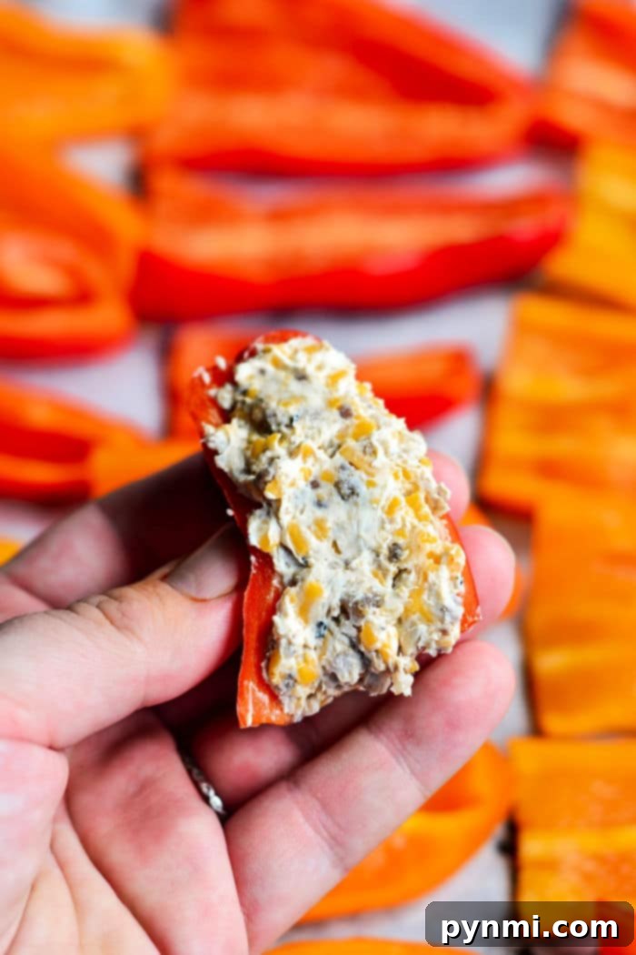 A hand holds a stuffed mini sweet pepper filled with a blend of cream cheese and herbs. Slices of red and orange peppers are blurred in the background, adding a colorful touch.