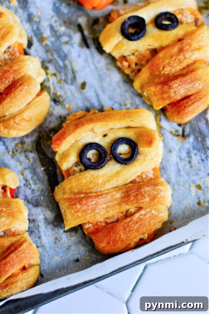 Freshly baked crescent roll mummies and stuffed mini sweet peppers rest on a parchment-lined baking sheet, the mummies shaped with olive slice eyes and wrapped dough for that classic bandage look.