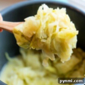 A wooden spoon holds a portion of buttered cabbage over a pot, cooked to softness in the pressure cooker. The cabbage appears lightly seasoned with a hint of butter, showcasing its pale green color. The pot is partially visible in the background.