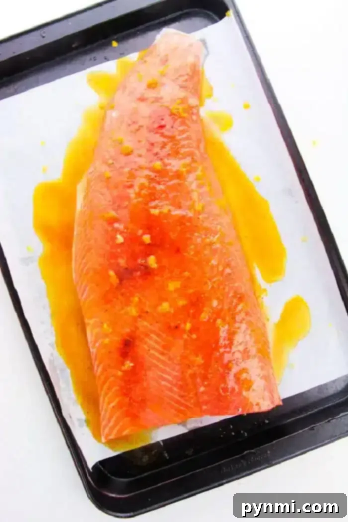 A marinated piece of raw salmon, placed on a baking sheet lined with parchment paper, adorned with fresh herbs and lemon slices, ready for the oven.
