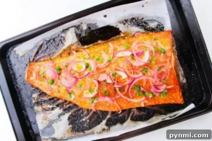A cooked salmon fillet lies on a parchment-lined baking tray. The salmon is garnished with thinly sliced red onions and chopped parsley. Wondering how long to bake salmon at 400? The tray shows some residue from the cooking process, indicating it was baked to perfection, ready to be served.