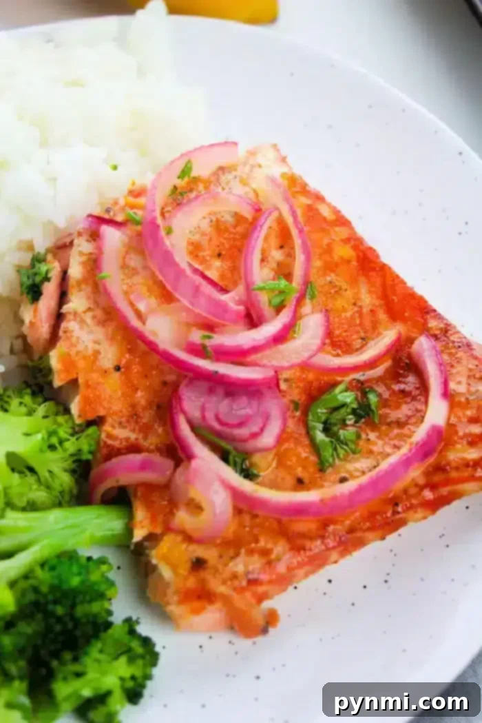 A close-up of a plate featuring a piece of succulent baked salmon topped with thinly sliced red onions and herbs. The plate also includes servings of fluffy white rice and vibrant steamed broccoli, creating a complete and balanced meal.