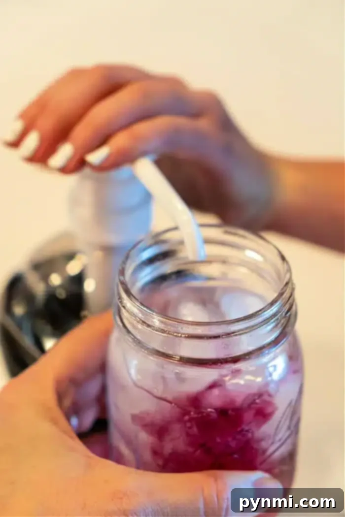 Craft Your Own Refreshing Lotus Drink 6 A person pumps liquid from a dispenser into a mason jar filled with a refreshing Lotus iced drink. The person's other hand carefully holds the jar while the pump's nozzle hovers above it, adding the perfect touch. In the softly blurred background, hints of vibrant colors dance subtly out of focus.