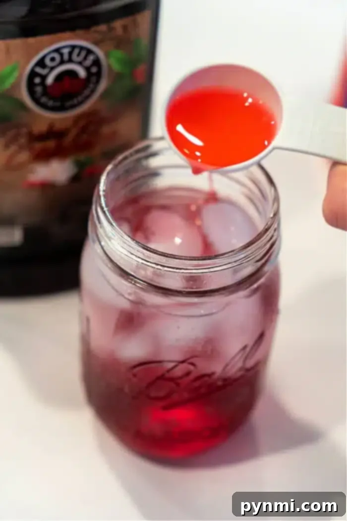 Craft Your Own Refreshing Lotus Drink 7 A measuring spoon pours red liquid into a Ball Mason jar packed with ice and another red liquid, creating a tantalizing Lotus Drink. A partially visible bottle labeled Lotus hints at the refreshing flavors waiting to be discovered.
