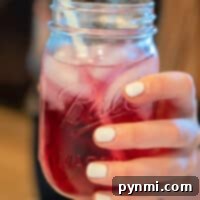 Craft Your Own Refreshing Lotus Drink 8 A hand holds a mason jar filled with a vibrant red Lotus drink, complete with ice cubes and a straw; nails are painted in crisp white.