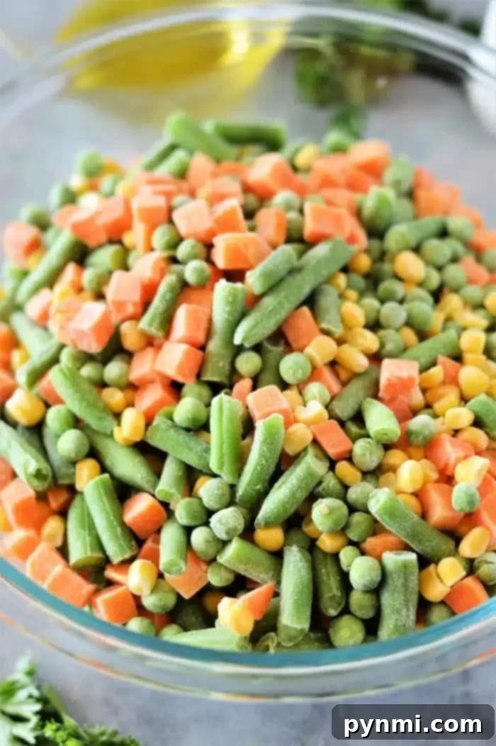 Frozen to Fantastic: Roasting Your Veggies 4 A clear glass bowl brimming with a colorful medley of frozen mixed vegetables—vibrant green beans, sweet peas, crunchy diced carrots, and golden corn kernels. The vegetables are ready for seasoning, sitting on a subtle gray countertop, with a hint of fresh parsley for garnish in the background.