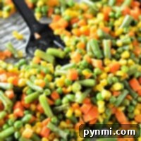 A close-up shot of perfectly roasted mixed vegetables—green beans, corn, peas, and carrots—arranged on a baking sheet, with a black serving spoon resting nearby, ready to scoop up the delicious, caramelized veggies.