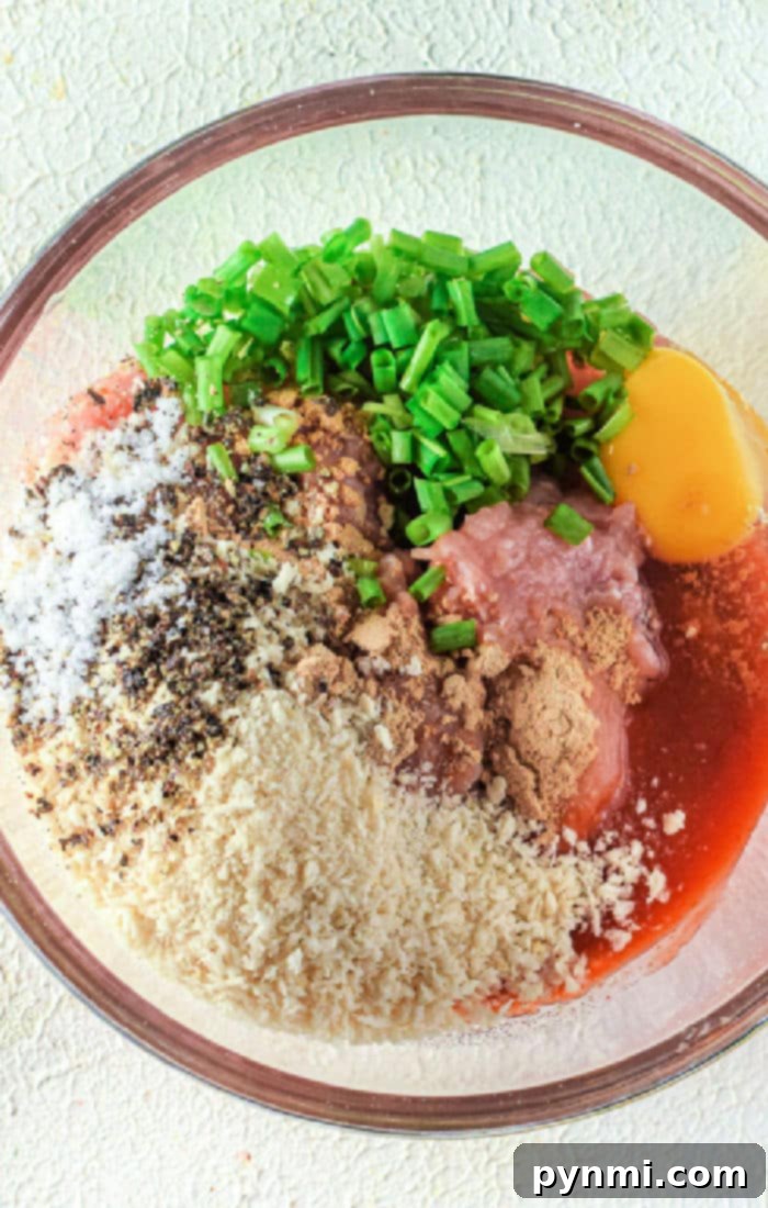 Crispy Air-Fried Chicken Meatballs 3 A glass bowl holds the preparation for Air Fryer Chicken Meatballs: raw minced meat, an egg, breadcrumbs, chopped green onions, black pepper, salt, ginger powder, and a drizzle of red sauce ready to be transformed into flavorful bites.