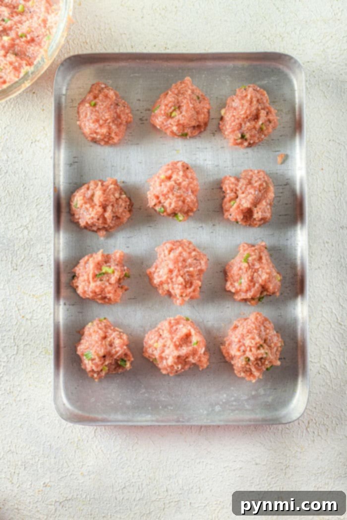 Crispy Air-Fried Chicken Meatballs 6 A metal baking tray holds twelve evenly spaced chicken meatballs, ready for the air fryer. The pink meatballs are flecked with visible herbs and spices, arranged in four neat rows. The tray rests on a textured, light-colored surface, promising a deliciously crisp finish.