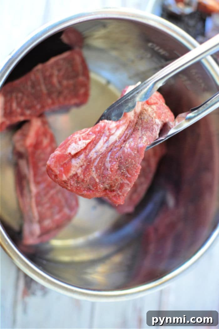 A pair of tongs holding raw beef over a metal pot, ready to transform them into flavorful Instant Pot Birria Tacos that are sure to delight.