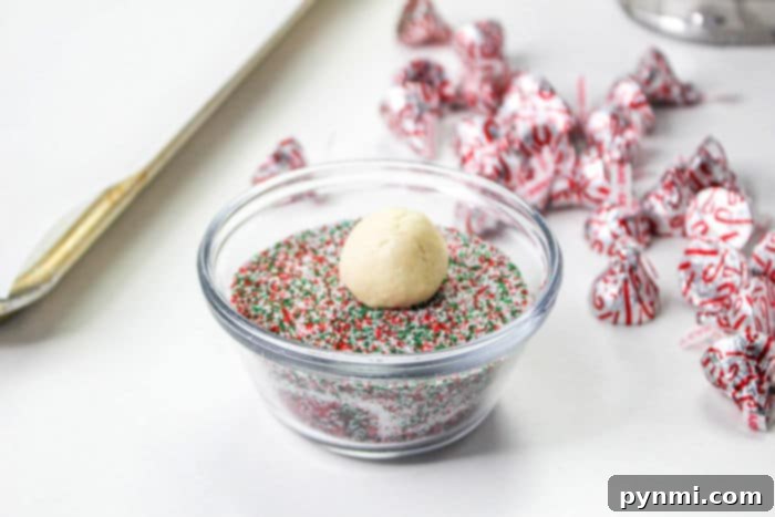 Blooming Bites 5 Four freshly baked peppermint thumbprint cookies arranged neatly on a plate, each featuring a classic Hershey Kiss in its center and dusted with festive red and green sprinkles. The cookies have a slightly cracked texture, indicating their delicious chewiness.