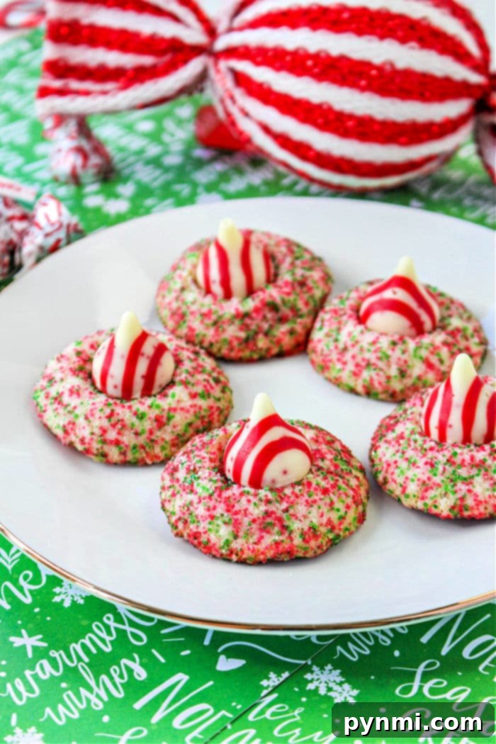 Blooming Bites 8 A festive holiday platter adorned with five vibrant Hershey Kiss Christmas Cookies. Each cookie is generously coated in red and green sprinkles and crowned with a red and white striped Hershey Kiss, adding a pop of color. In the background, a large, decoratively wrapped present in matching red and white stripes with a green base enhances the festive theme.