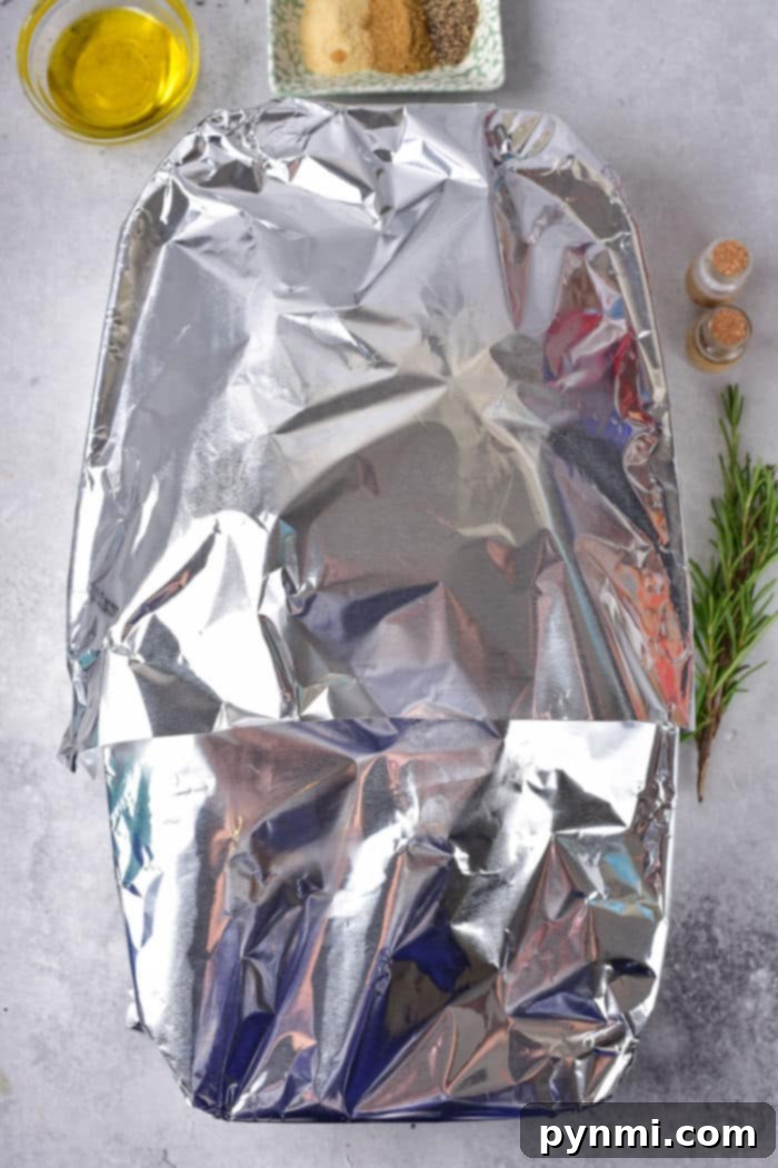 A frozen duck nestled in a baking dish, covered with aluminum foil, centered on a marble countertop. Alongside are a small bowl of olive oil, a plate with seasoning, two small shakers, and a sprig of fresh rosemary, awaiting the cooking process.