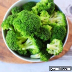A bowl brimming with vibrant green broccoli florets sits on a wooden and white surface, ready to be transformed into a quick microwave broccoli masterpiece.