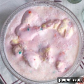 Pink Delight Punch 6 A bowl of pink ice cream punch, inspired by the ultimate Pink Punch Recipe, is adorned with colorful sprinkles on a wooden table. Beside it lies a striped napkin and silver spoons, ready for indulgence.