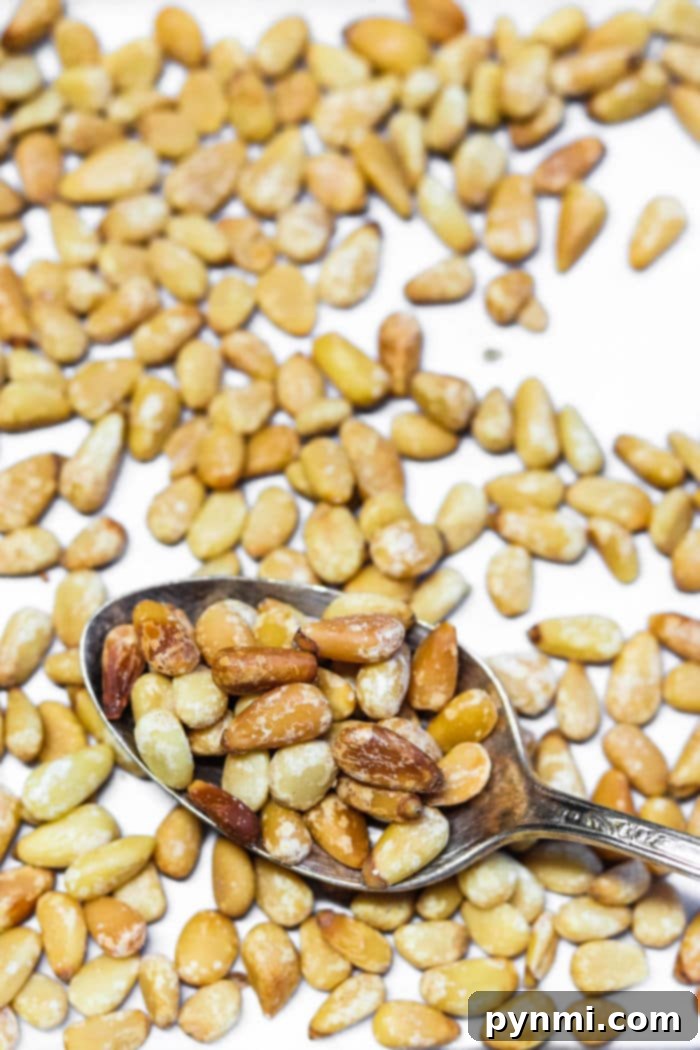Close-up of a spoon filled with perfectly roasted pine nuts scattered on a white surface, showcasing the simple elegance of how to roast pine nuts to golden perfection.