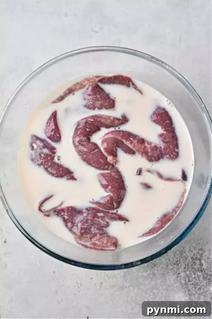 Tender Pan-Fried Beef Liver with Caramelized Onions 4 A glass bowl filled with pieces of raw liver soaking in milk rests on a light-colored, textured surface, illustrating the soaking process for beef liver.