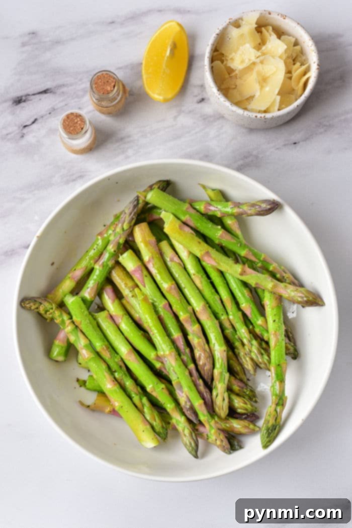 A bowl of asparagus spears with a lemon wedge, shaved cheese, and spices rests on a marble surface, perfect for those wondering how long to cook asparagus in the oven for a fragrant delight.