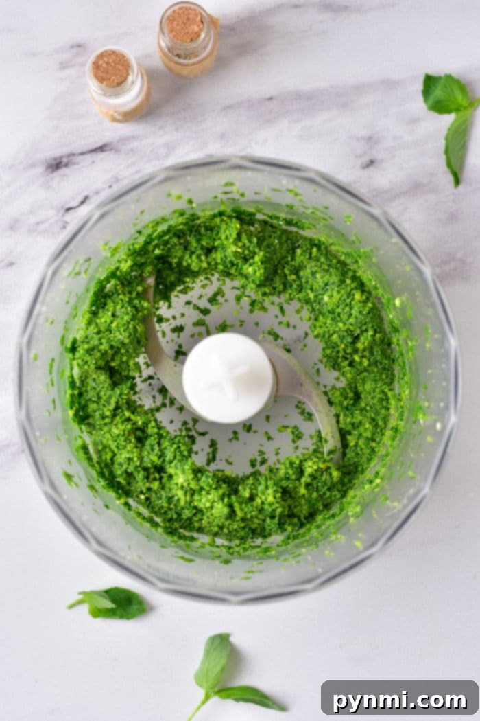 Effortless Homemade Pine Nut Pesto 4 Green pesto without pine nuts swirls in a food processor bowl on a marble countertop, accompanied by fresh basil leaves and spice jars nearby.