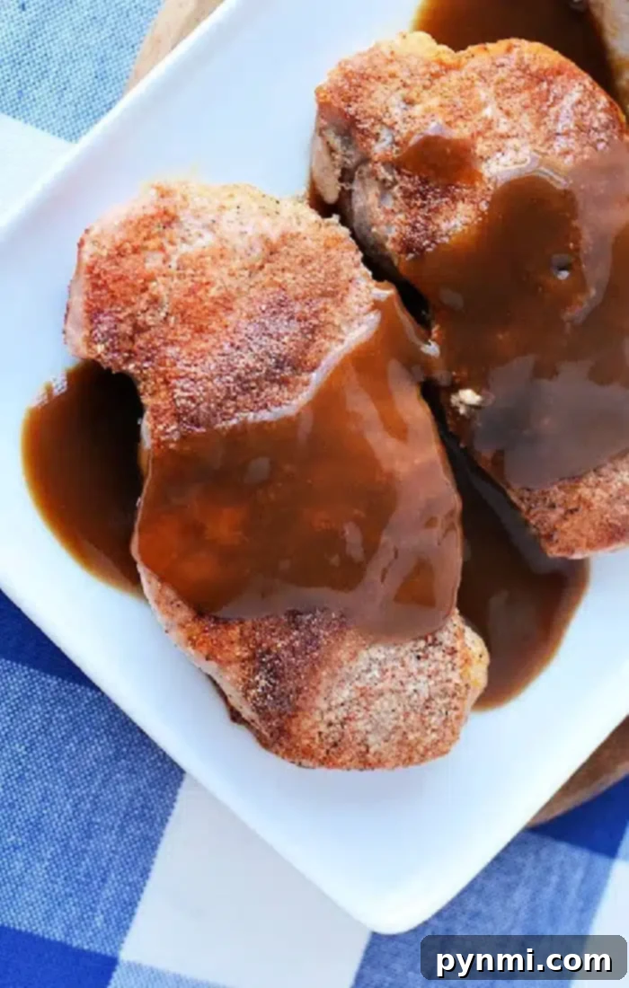 Two cooked pork chops with gravy sit on a white plate, the result of expertly cooking frozen pork chops. The plate rests on a surface adorned with a blue and white checked pattern.