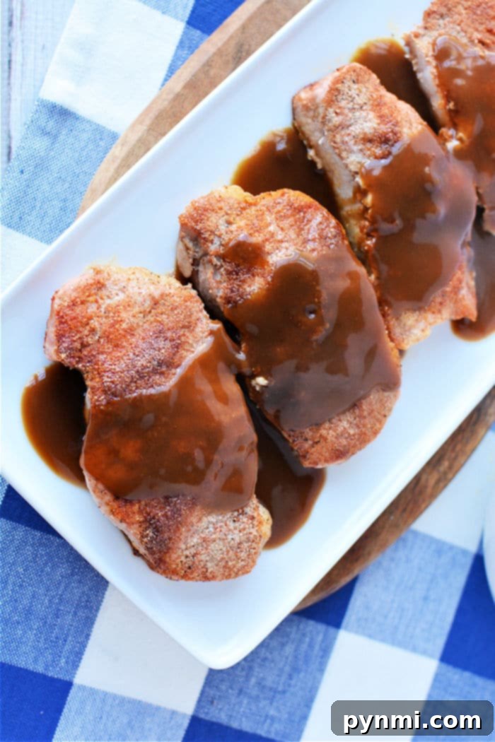 Four pieces of seasoned pork chops, expertly cooked from frozen, are topped with rich brown gravy and arranged on a rectangular white plate. The dish is beautifully set on a wooden board over a blue and white checkered tablecloth.