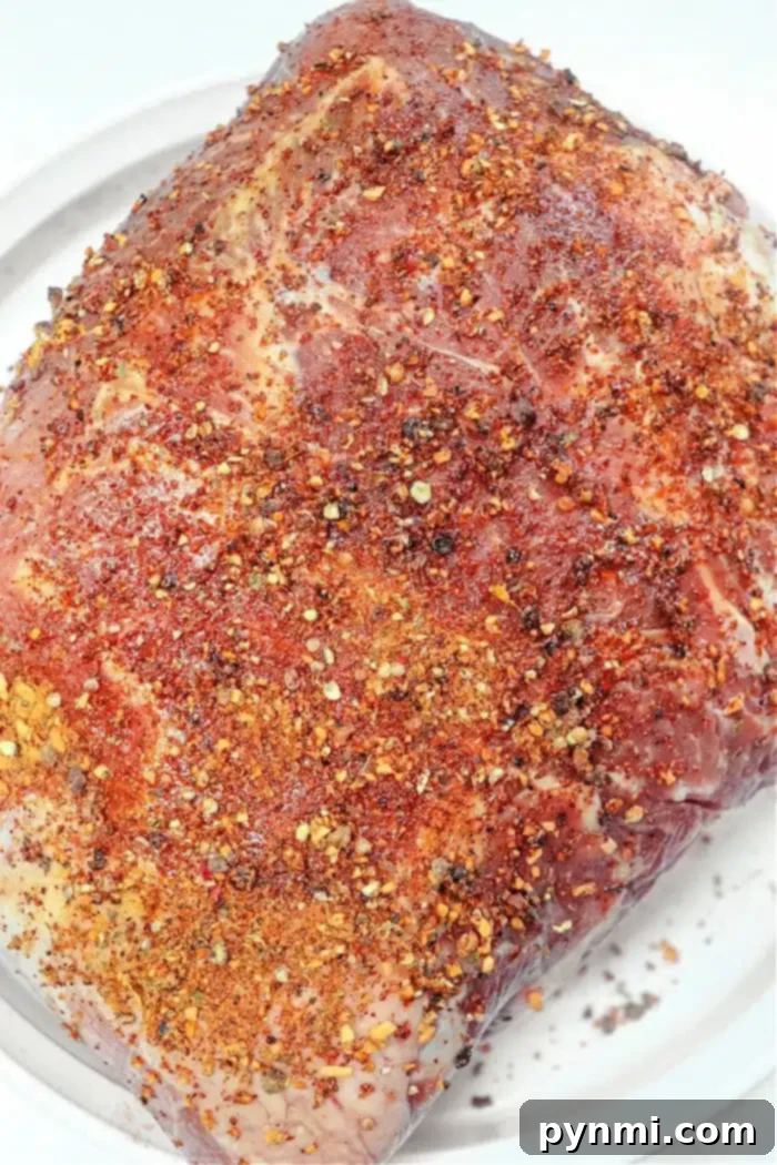 Slow-Smoked Pork Perfection 3 A raw pork butt generously coated in a vibrant dry rub, featuring visible red pepper flakes, ground pepper, and various fragrant herbs. The meat is ready for smoking, promising a rich and flavorful outcome.