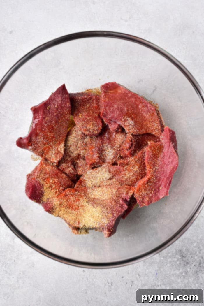 Mastering Beef Heart: Tender Recipes and Techniques 4 A glass bowl containing raw beef heart pieces all coated with various seasonings, is placed on a light gray surface.