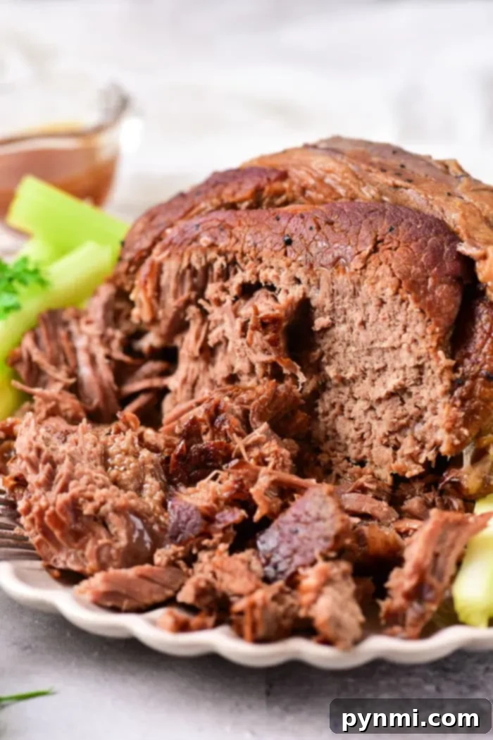 Tender Oven Roasted Beef Shoulder 2 Close-up of a cooked beef roast shoulder on a white plate, surrounded by celery pieces. The roast is tender and slightly shredded, with a visible moist texture. A small bowl with sauce is in the background, perfectly complementing this delicious dish.