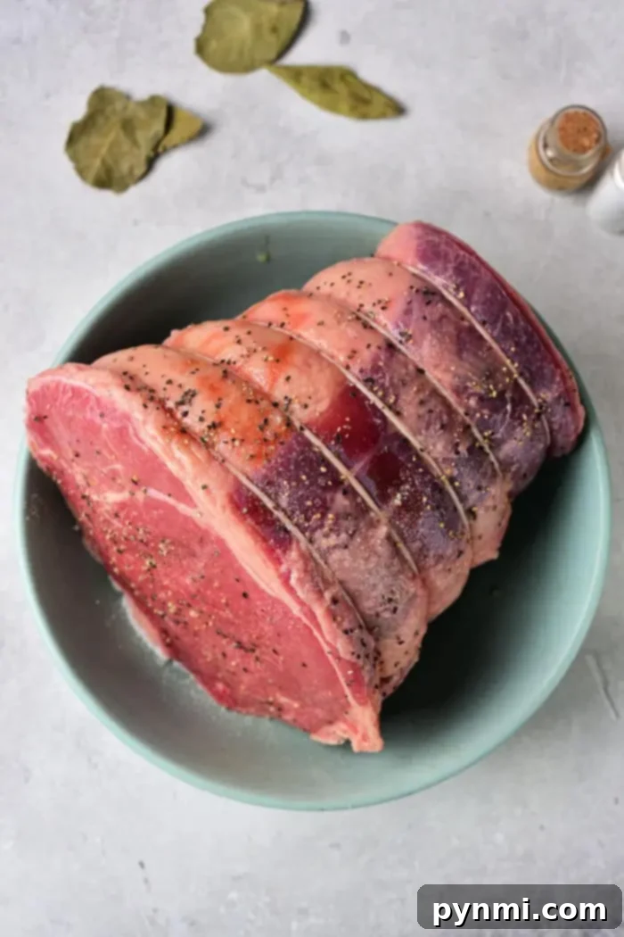 Tender Oven Roasted Beef Shoulder 3 A raw, tied beef roast shoulder, seasoned with salt and pepper, sits in a light green bowl on a gray surface. A few bay leaves and a small salt shaker add to the scene in the background.