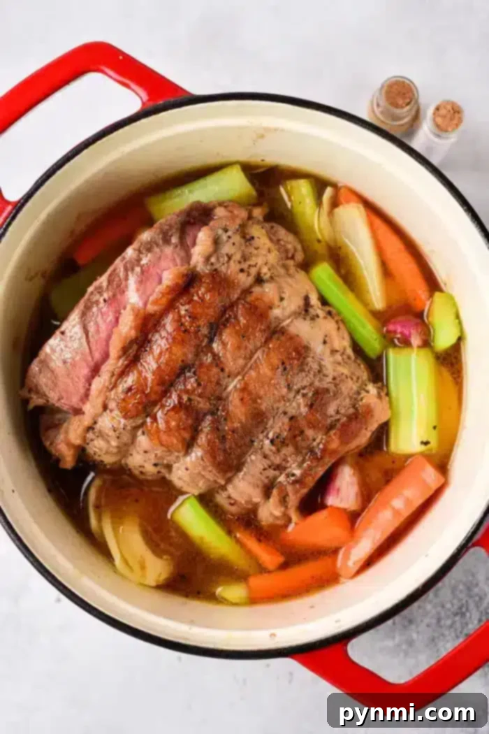 Tender Oven Roasted Beef Shoulder 4 A beef roast is cooking in a red cast iron pot, surrounded by various vegetables including carrots, celery, and onions, all simmering in a rich brown broth. The tender shoulder cut enhances the flavor perfectly. Two small spice containers are visible in the background.