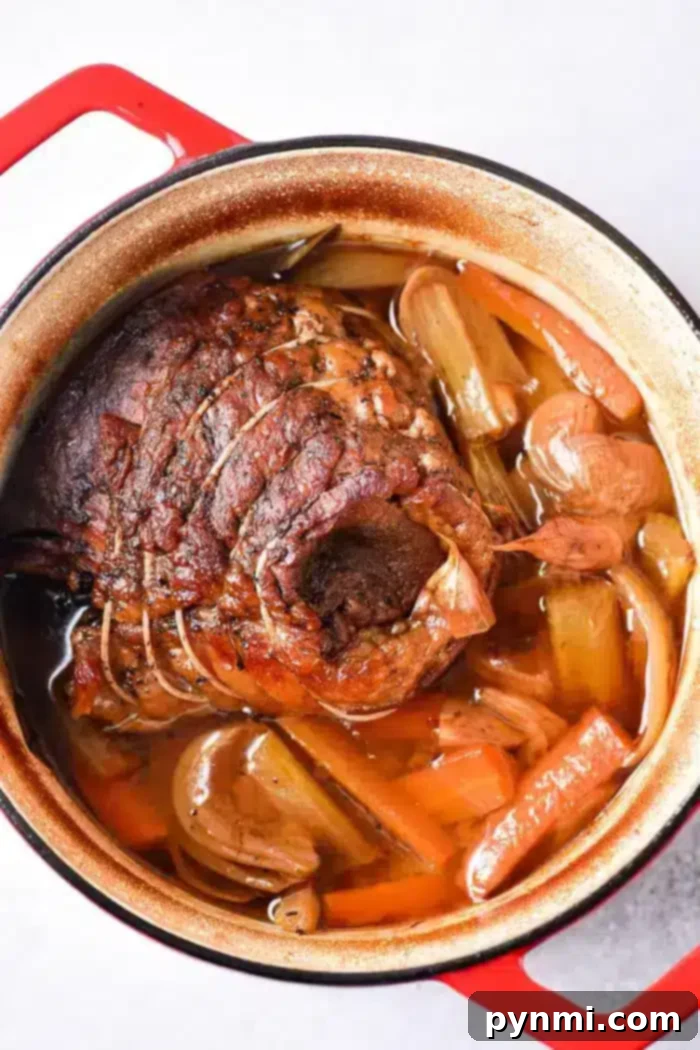 Tender Oven Roasted Beef Shoulder 5 A cooked beef roast shoulder in a red Dutch oven, surrounded by chunks of carrots, celery, and onions in a brown broth. The meat is seasoned and tied with string, resting in a savory liquid with visible spices.