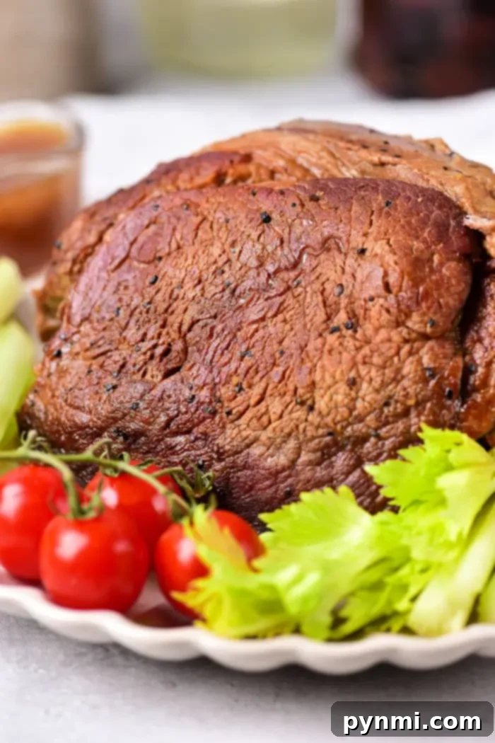 Tender Oven Roasted Beef Shoulder 7 A roasted beef roast shoulder served on a white plate, garnished with fresh cherry tomatoes and celery stalks. The beef appears well-seasoned with a brown crust. A blurred bowl of sauce is visible in the background.