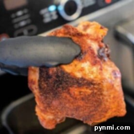 A gloved hand holds seasoned cooked turkey thigh, with an air fryer in the background, ready to also transform turkey thighs into a crispy delight.