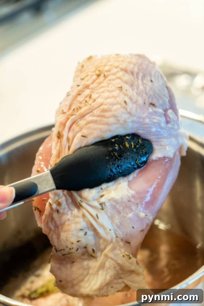 Close-up of a brined turkey breast being carefully lifted from a liquid-filled pot with black tongs, showing the meat glistening and ready for the next stage of cooking. The background is a softly blurred kitchen setting.