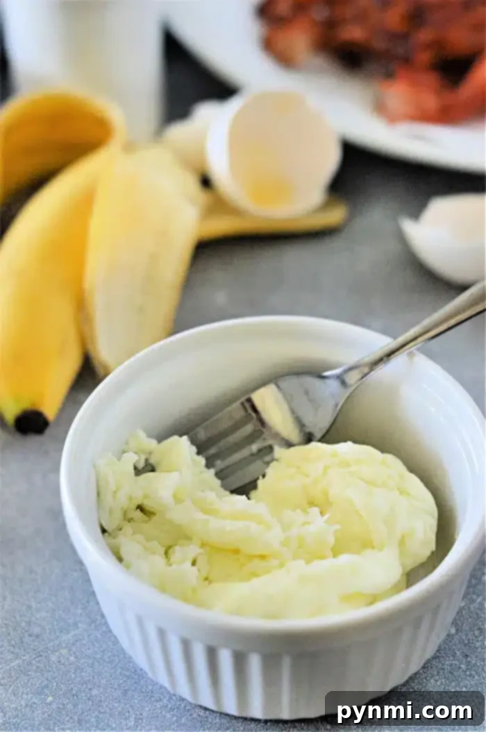 Perfectly Cooked Egg Whites Every Time 5 A small white bowl contains cooked egg whites with a fork resting inside. In the background, there are two unpeeled bananas, an empty eggshell from cooked egg whites, and a plate of crispy bacon on a gray surface, suggesting a full breakfast.