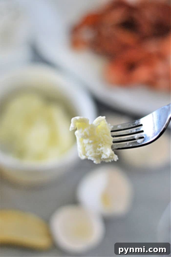 Perfectly Cooked Egg Whites Every Time 6 A close-up of a fork holding a small piece of cooked egg whites. In the blurred background, a white bowl filled with more cheese sits beside a plate of sliced meat and garlic cloves.