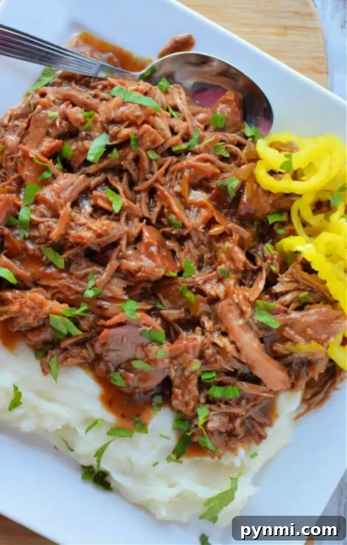 A plate of shredded Mississippi Pork Roast served over creamy mashed potatoes and garnished with fresh chopped parsley. A side of vibrant yellow pickled pepperoncinis adds a tangy contrast, and a spoon rests gracefully on the plate, ready for a delicious bite.