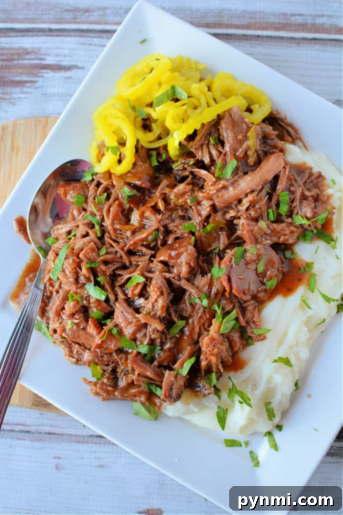 A beautifully plated dish featuring shredded Mississippi Pork Roast with rich gravy, artfully garnished with chopped herbs, alongside a creamy mound of mashed potatoes. Slices of bright yellow pepperoncini peppers provide a colorful and tangy accent, completing this satisfying meal.